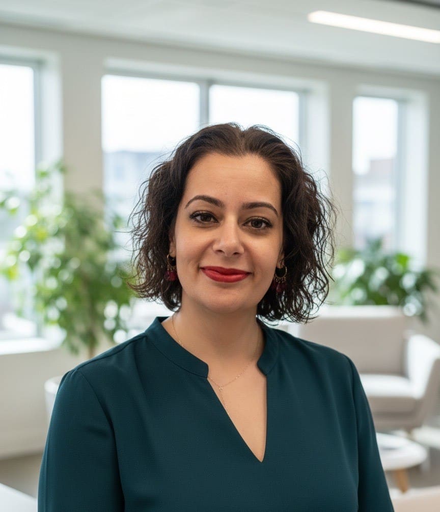 Woman with curly dark hair and red lipstick, wearing a teal blouse, smiles in a bright office with large windows and green plants in the background, creating a welcoming atmosphere perfect for coaching sessions.