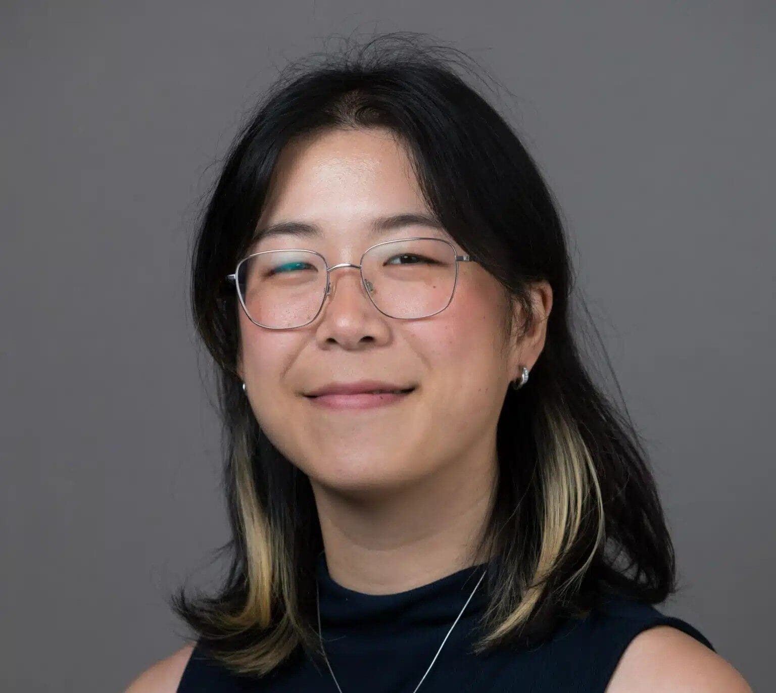 A person with shoulder-length dark hair with light tips, wearing glasses, a sleeveless dark top, and a necklace, smiles softly against a plain gray background—perfect for featuring in client testimonials.