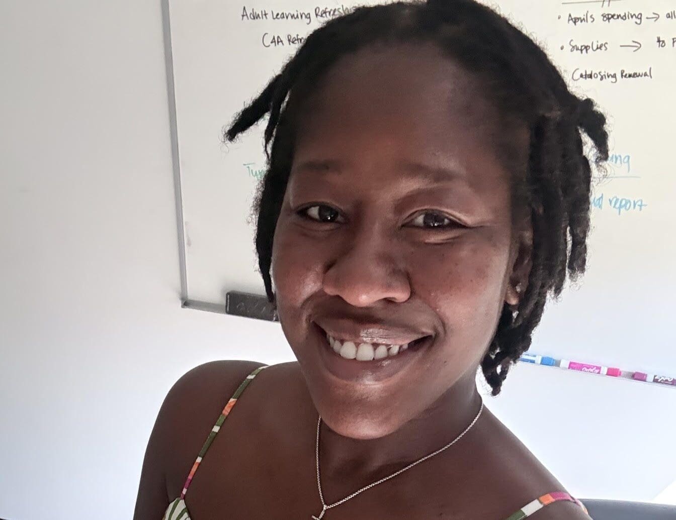 A woman with short, twisted hair smiles at the camera, wearing a colorful, patterned dress and a cross necklace. A whiteboard with notes and client testimonials is visible behind her in an office setting.